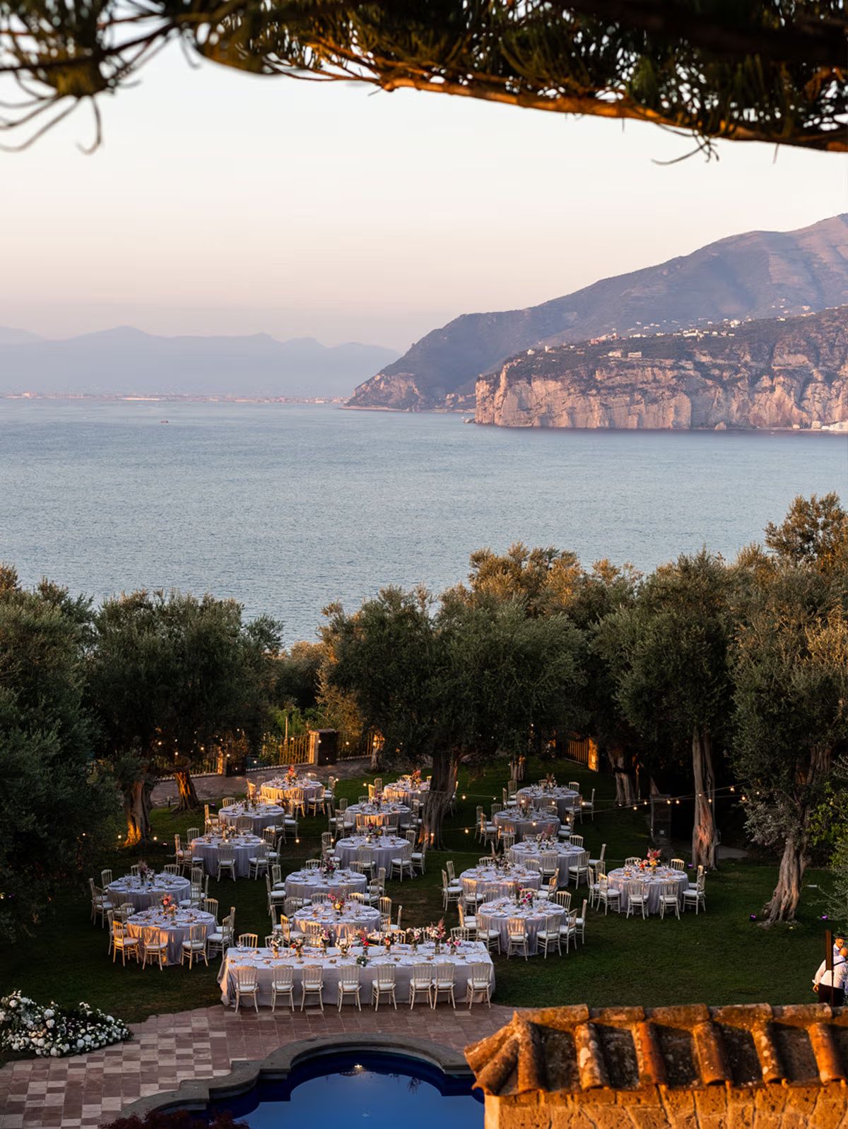 Dinner tables set among olive trees at sunset, with the Sorrento coastline and the sea behind